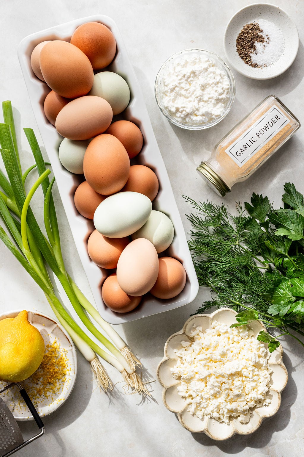 A tray of brown and pale eggs, green onions, a lemon, fresh herbs, grated cheese, cottage cheese, garlic powder, and a small dish of salt and pepper on a white surface.