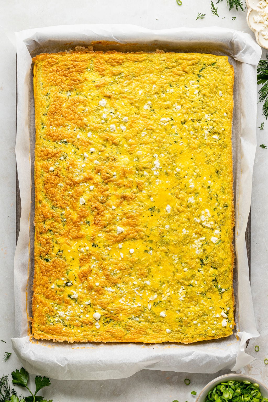 A large rectangular baked egg casserole with a golden top, garnished with small bits of white cheese and herbs, on parchment paper in a baking tray.