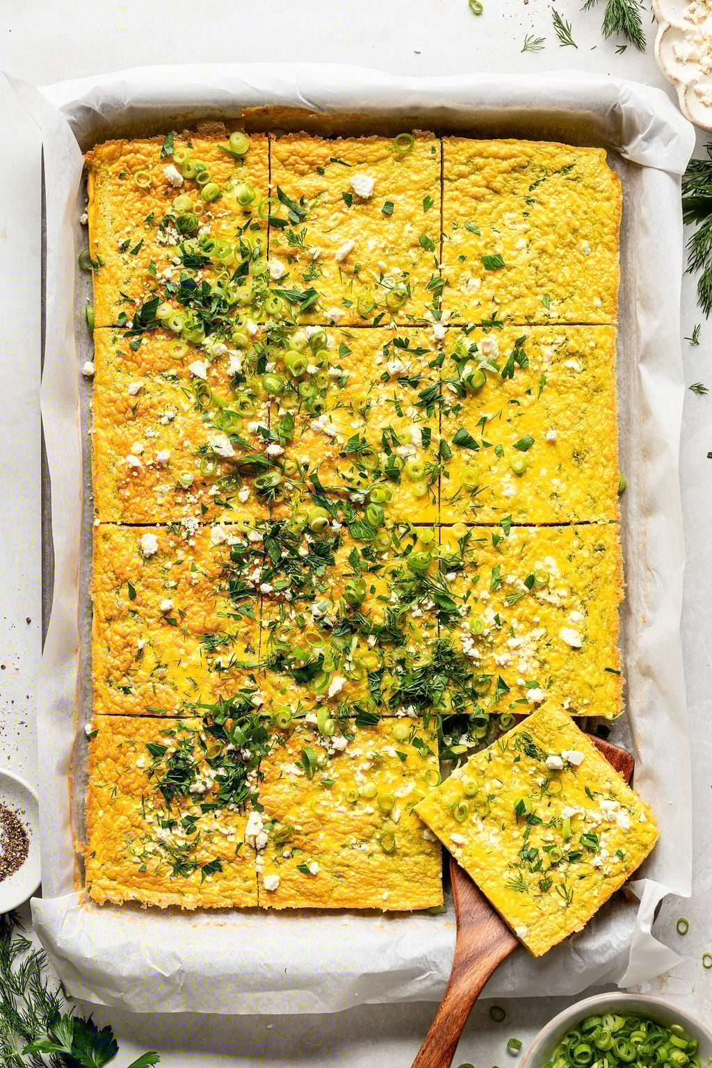 A rectangular baked egg casserole topped with chopped herbs, green onions, and crumbled cheese, cut into squares with one piece being served on a wooden spatula.