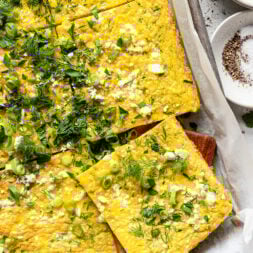 A sheet pan frittata topped with chopped herbs, green onions, and crumbled cheese, cut into squares and served on parchment paper.