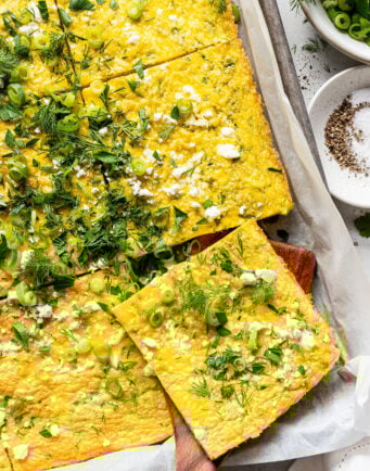 A sheet pan frittata topped with chopped herbs, green onions, and crumbled cheese, cut into squares and served on parchment paper.