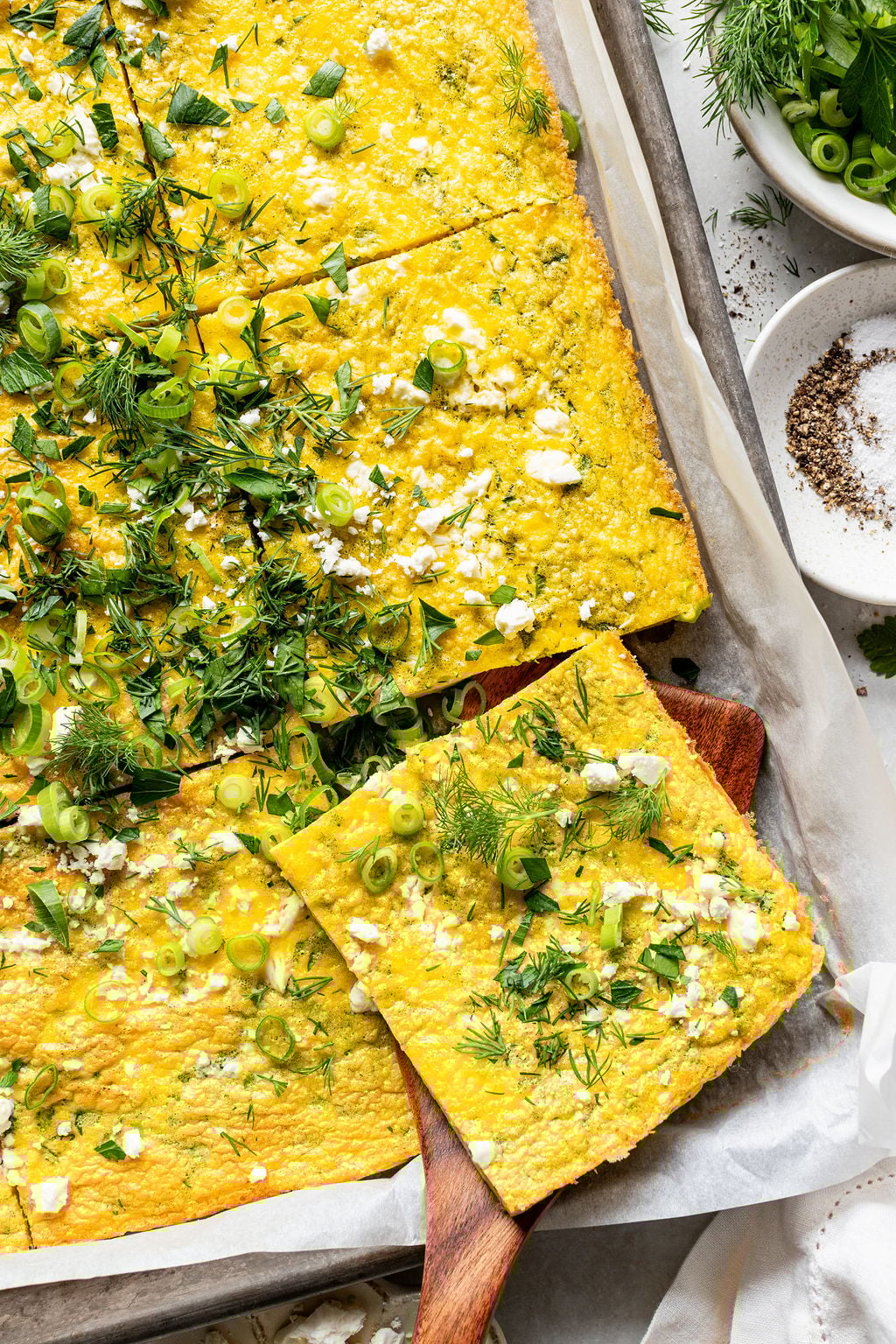 A sheet pan frittata topped with chopped herbs, green onions, and crumbled cheese, cut into squares and served on parchment paper.
