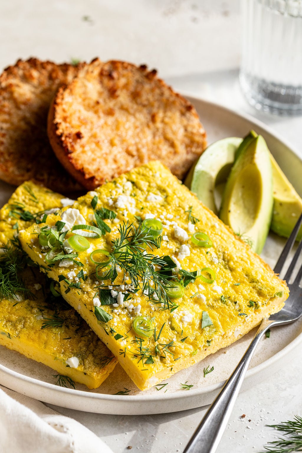 A plate with two rectangular pieces of baked egg topped with herbs and cheese, sliced avocado, and toasted English muffins in the background.