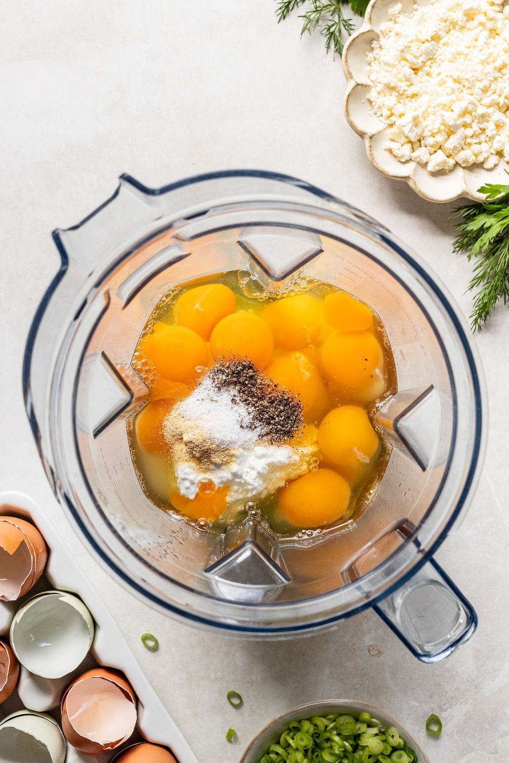 A blender filled with raw eggs, spices, and seasoning on a countertop, surrounded by eggshells, chopped green onions, and crumbled cheese.