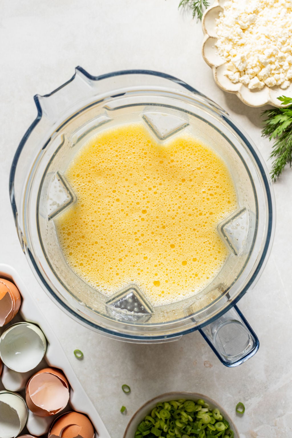 A blender filled with a frothy yellow egg mixture sits on a counter next to cracked egg shells, chopped green onions, dill, and crumbled cheese.