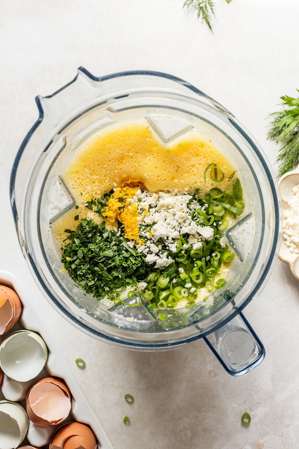 A blender containing a mixture of beaten eggs, chopped herbs, green onions, crumbled cheese, and lemon zest, with empty eggshells nearby.