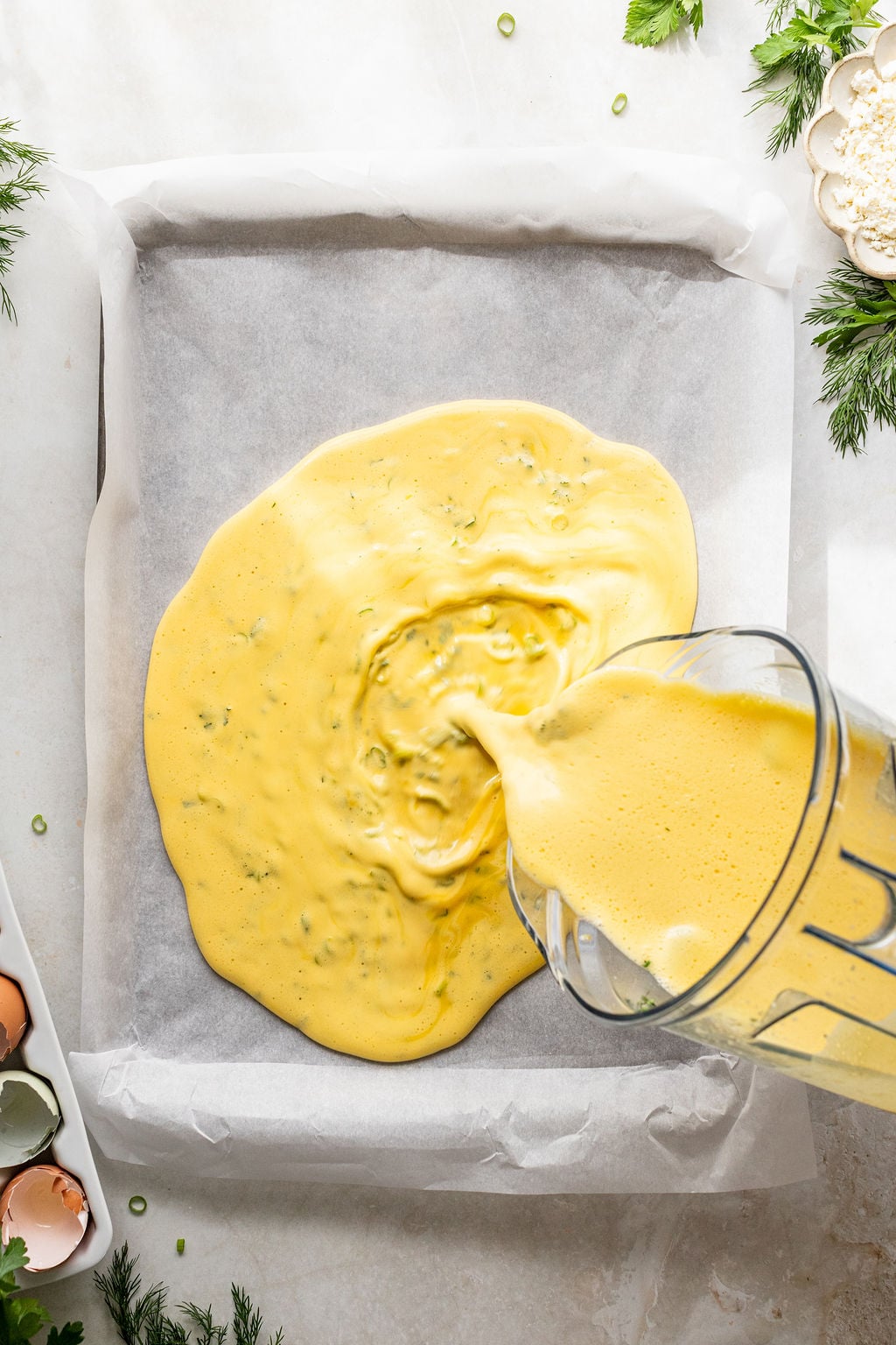 A glass pitcher pours a yellow egg mixture into a parchment-lined baking dish on a white surface, surrounded by herbs, eggshells, and a bowl of flour.