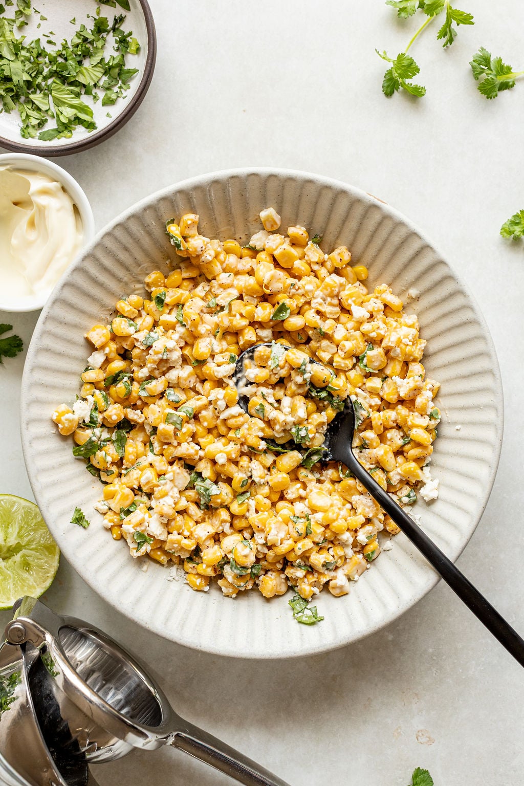 A bowl of Mexican street corn salad with a spoon, garnished with cilantro and crumbled cheese. Surrounding the bowl are chopped herbs, lime, and a citrus press on a white surface.