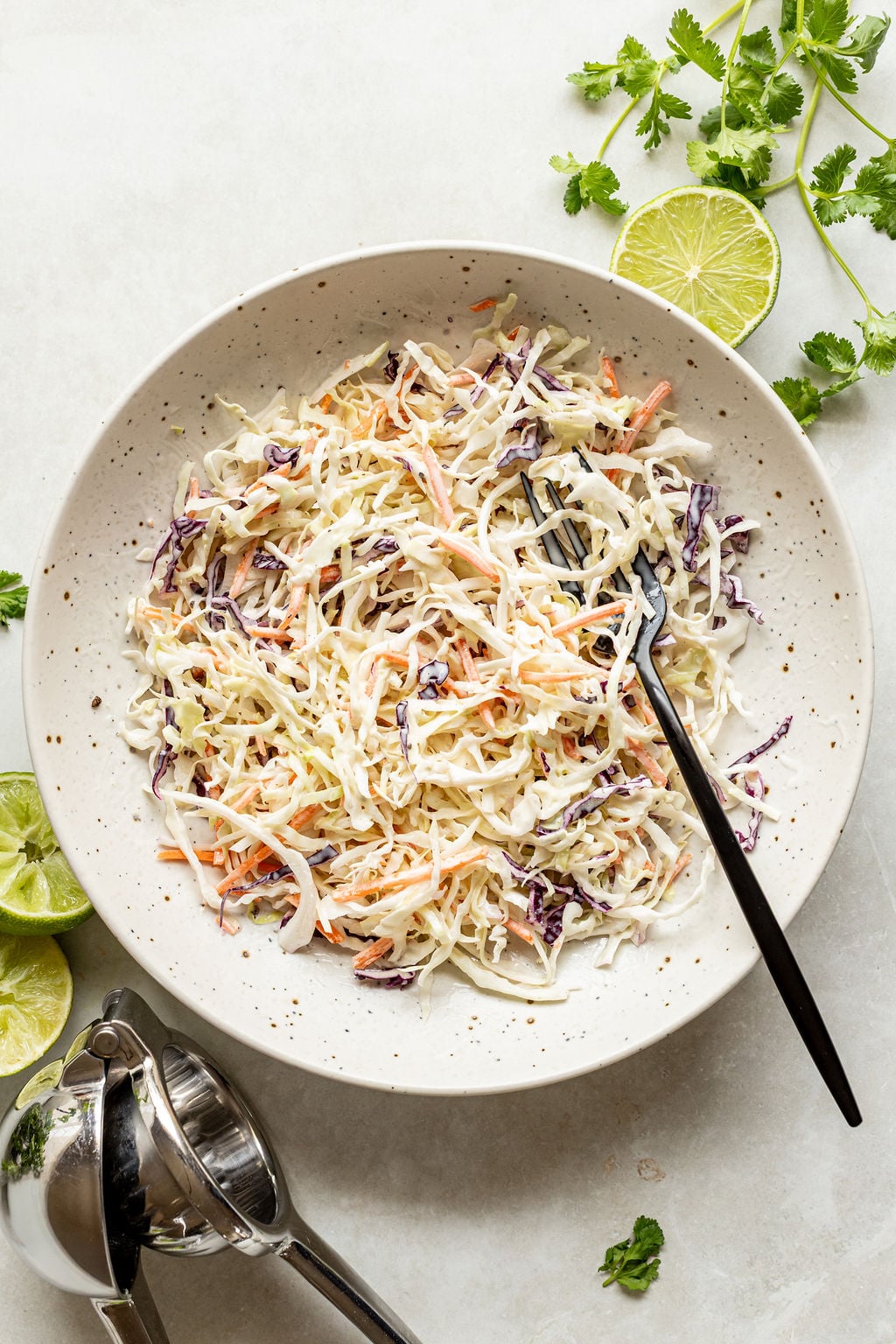 A bowl of coleslaw with shredded cabbage and carrots sits on a white surface, accompanied by a fork, cilantro, lime halves, and a metal citrus juicer.