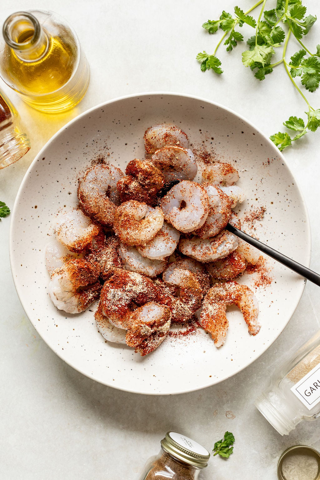 Raw shrimp in a white bowl, sprinkled with spices, with a black spoon. Olive oil, garlic powder, herbs, and spice jars are nearby on a white surface.