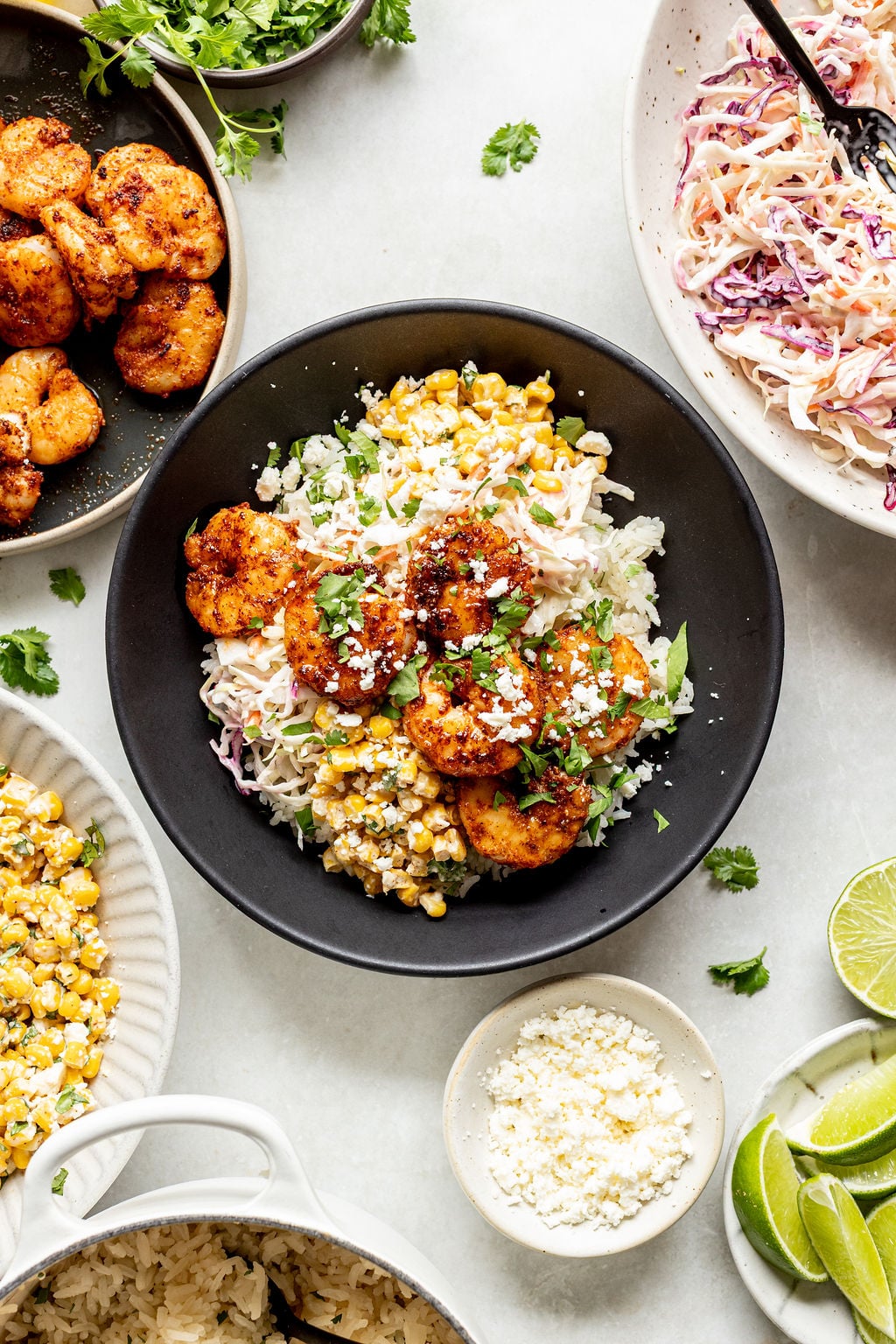 A black bowl filled with rice, grilled shrimp, corn, slaw, and cheese, surrounded by plates of shrimp, corn, slaw, lime wedges, and cheese on a white surface.