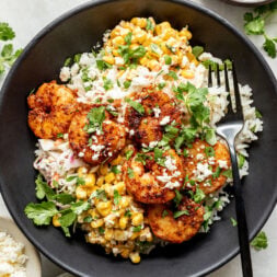 A black bowl with grilled shrimp, cilantro rice, street corn, crumbled cheese, and fresh cilantro, served with lime wedges and a fork.