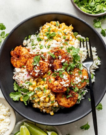 A black bowl with grilled shrimp, cilantro rice, street corn, crumbled cheese, and fresh cilantro, served with lime wedges and a fork.
