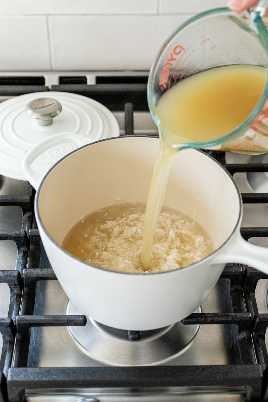 A hand pours broth from a measuring cup into a white pot containing rice on a stove.