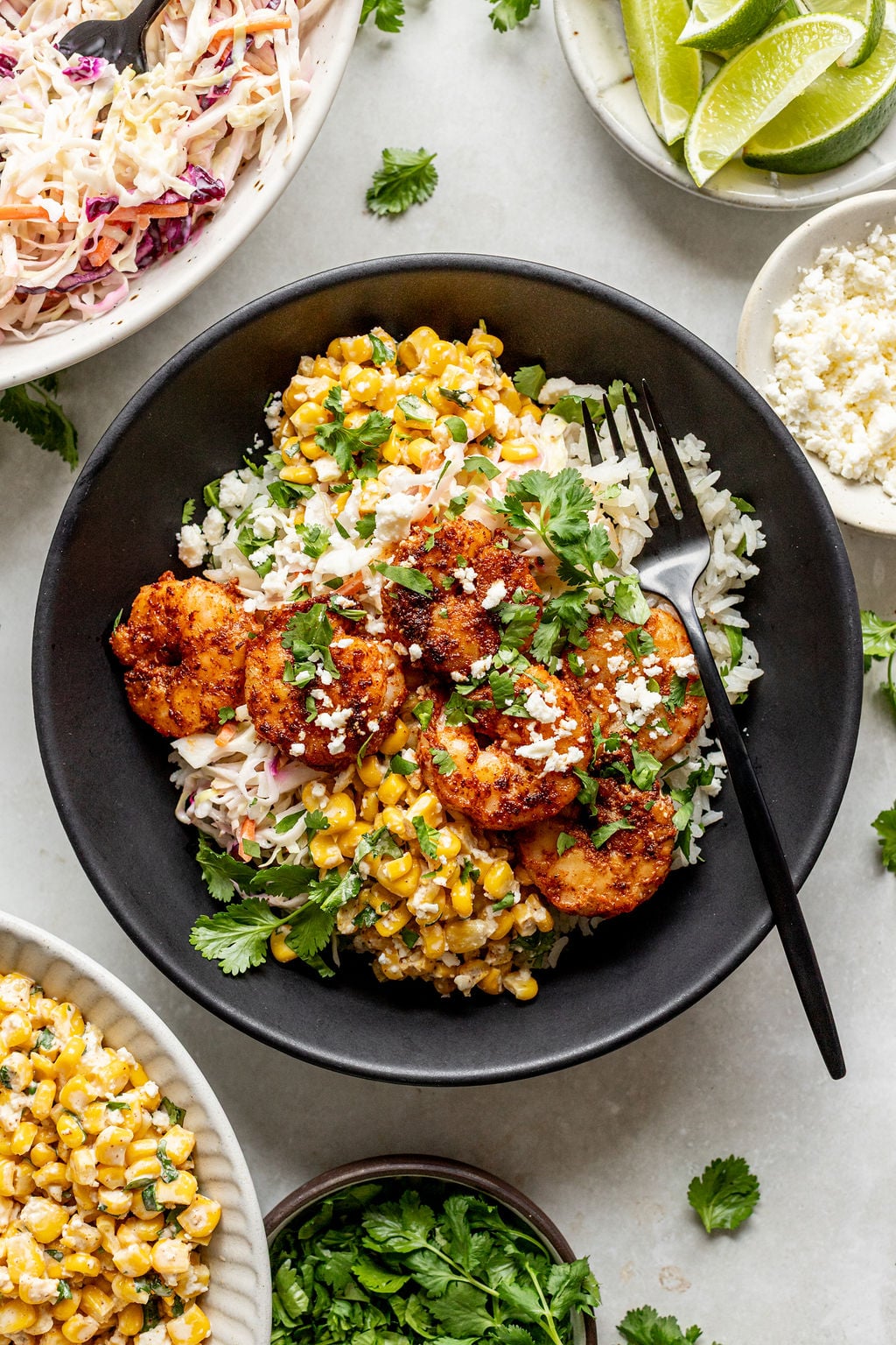 A bowl of rice topped with cooked shrimp, corn, slaw, cilantro, and crumbled cheese, with a black fork resting inside. Lime wedges, more corn, slaw, and cilantro are nearby.