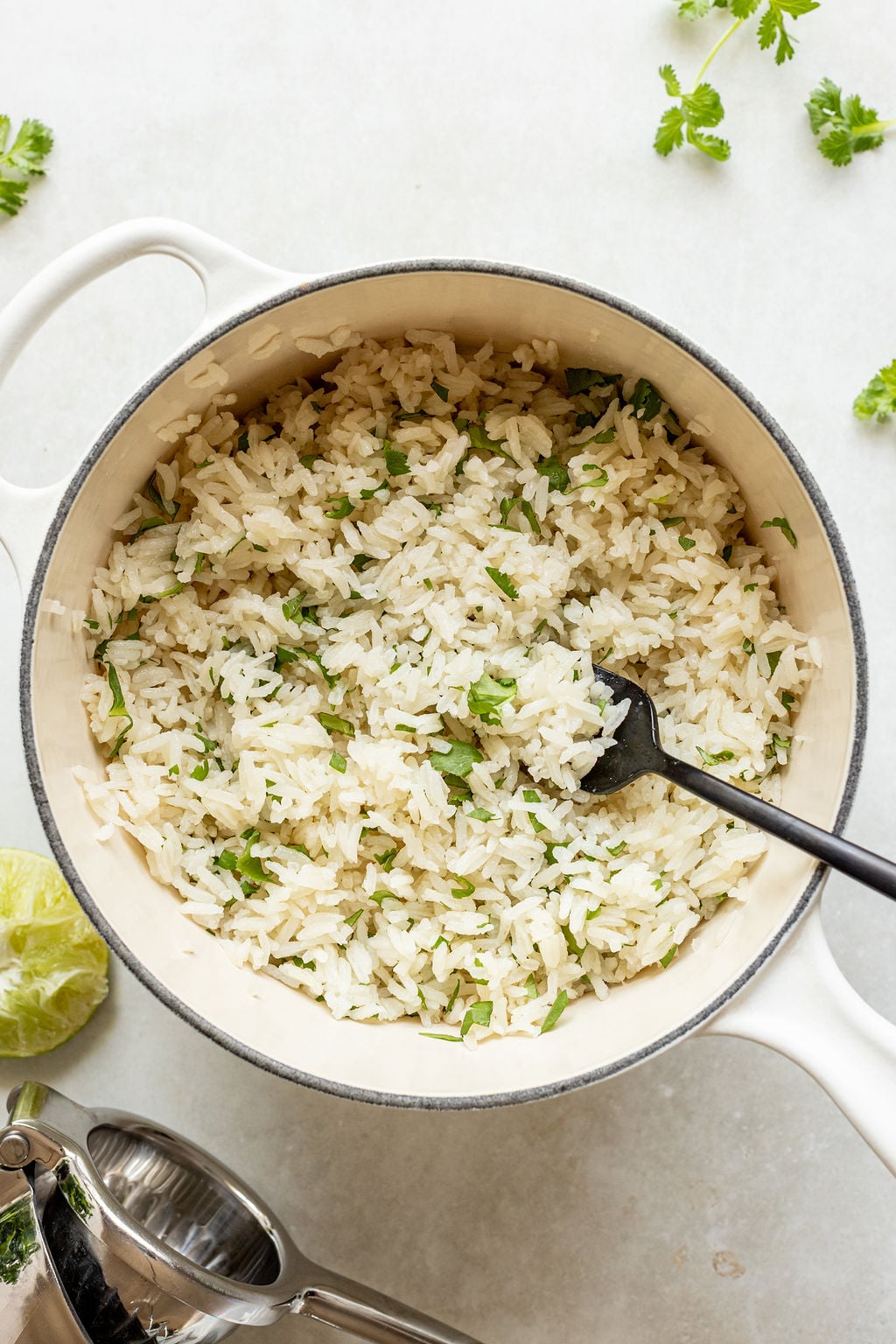 Cooked white rice mixed with chopped cilantro in a white pot, with a black serving spoon, lime and cilantro leaves nearby.