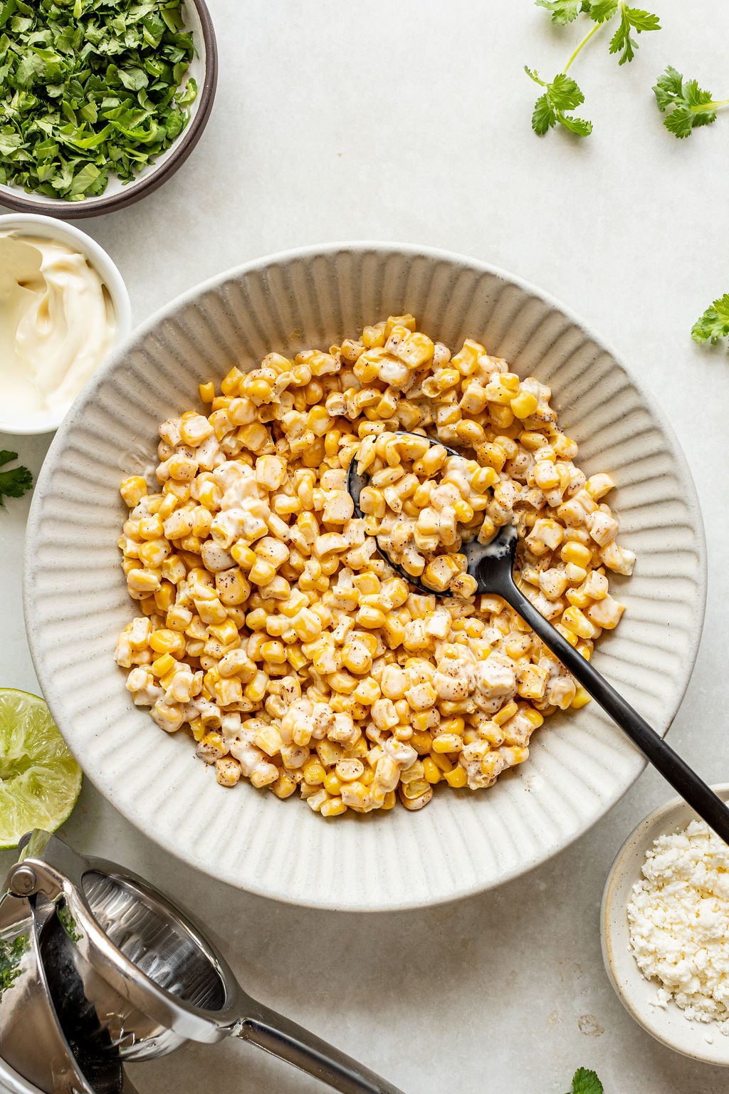 A white bowl filled with creamy seasoned corn kernels sits on a table, surrounded by small bowls of chopped herbs, mayonnaise, cheese, and a lime squeezer.