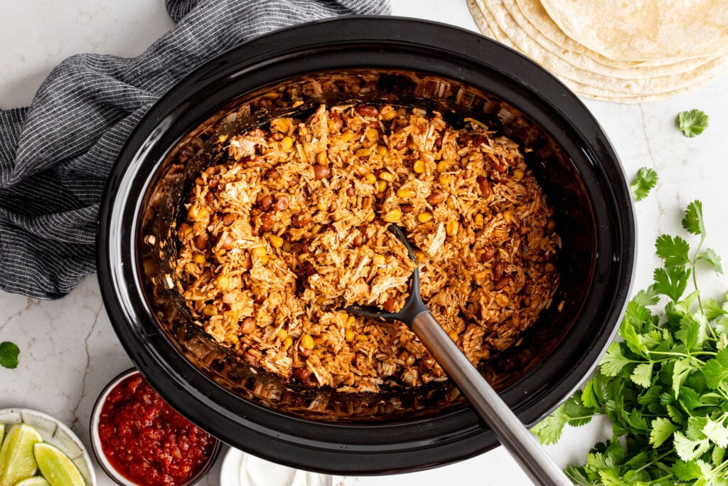 Shredded chicken, beans, and corn mixed together in a black slow cooker, with a serving spoon. Tortillas, salsa, lime wedges, sour cream, and cilantro are arranged around it.