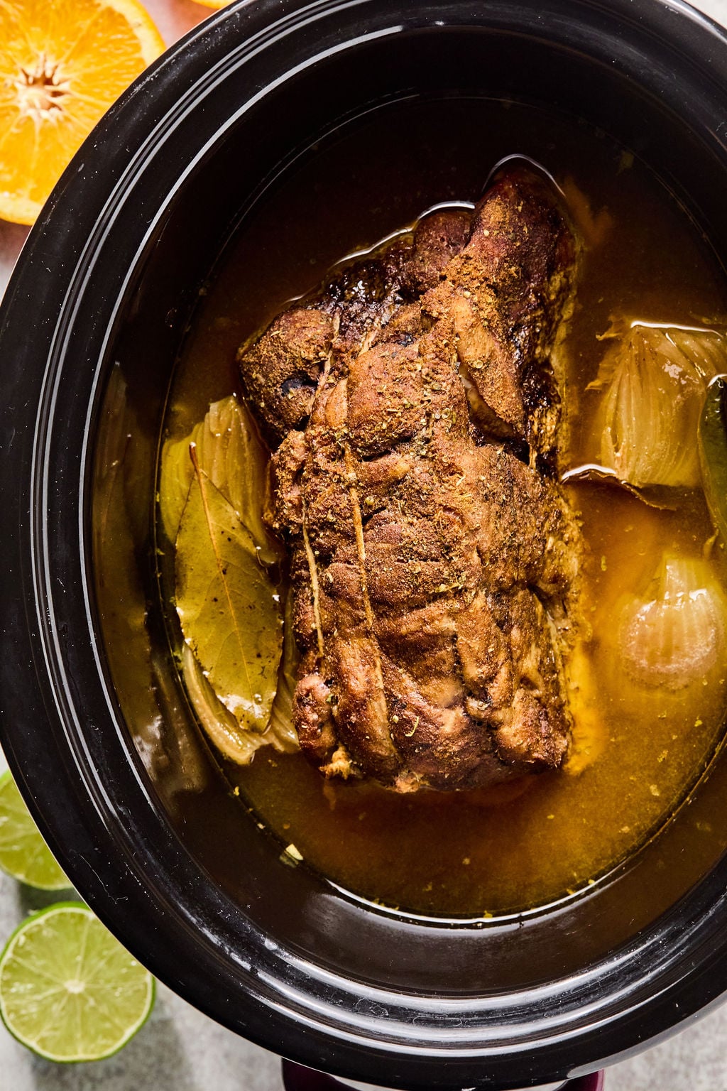 A seasoned pork roast cooks in broth with onions and bay leaves inside a slow cooker, surrounded by slices of lime and orange.