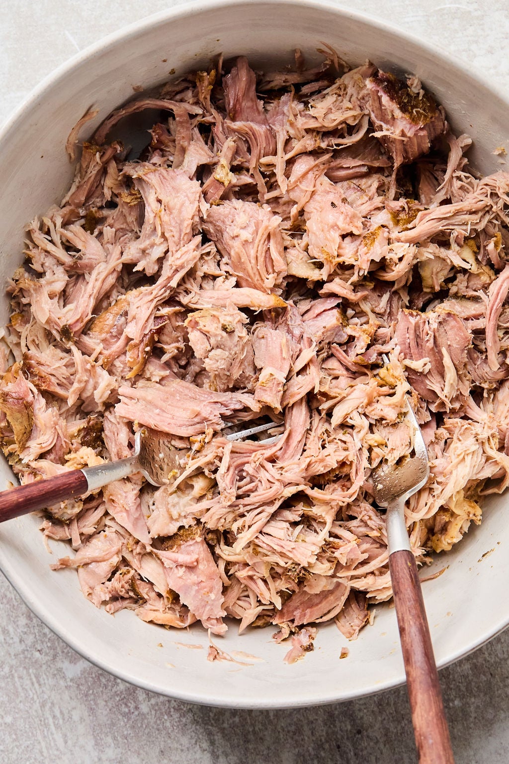 A bowl of shredded cooked pork with two forks, set on a light surface.