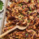 Shredded, crispy carnitas on a baking sheet with a wooden spoon, garnished with chopped fresh herbs.
