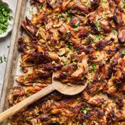 Shredded, crispy carnitas on a baking sheet with a wooden spoon, garnished with chopped fresh herbs.