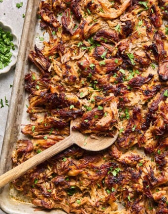 Shredded, crispy carnitas on a baking sheet with a wooden spoon, garnished with chopped fresh herbs.