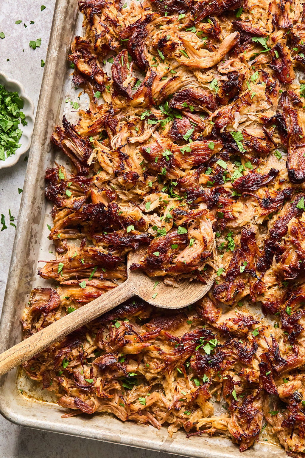Shredded, crispy carnitas on a baking sheet with a wooden spoon, garnished with chopped fresh herbs.