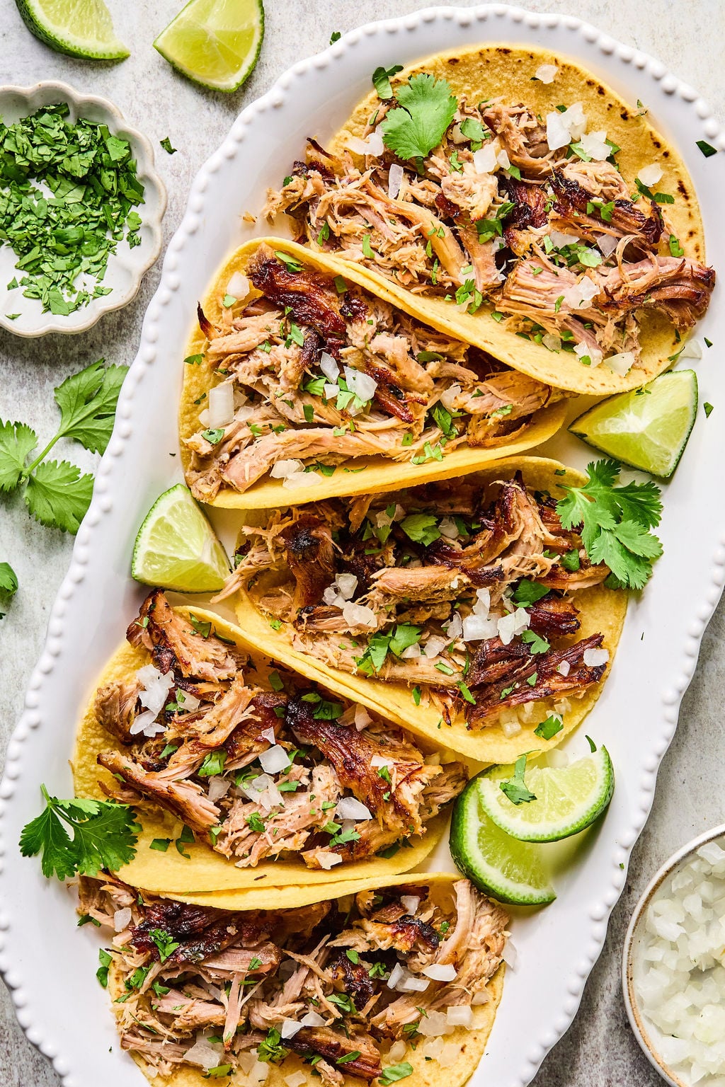Five shredded meat tacos in corn tortillas, garnished with chopped onions, cilantro, and lime wedges, arranged on a white platter.