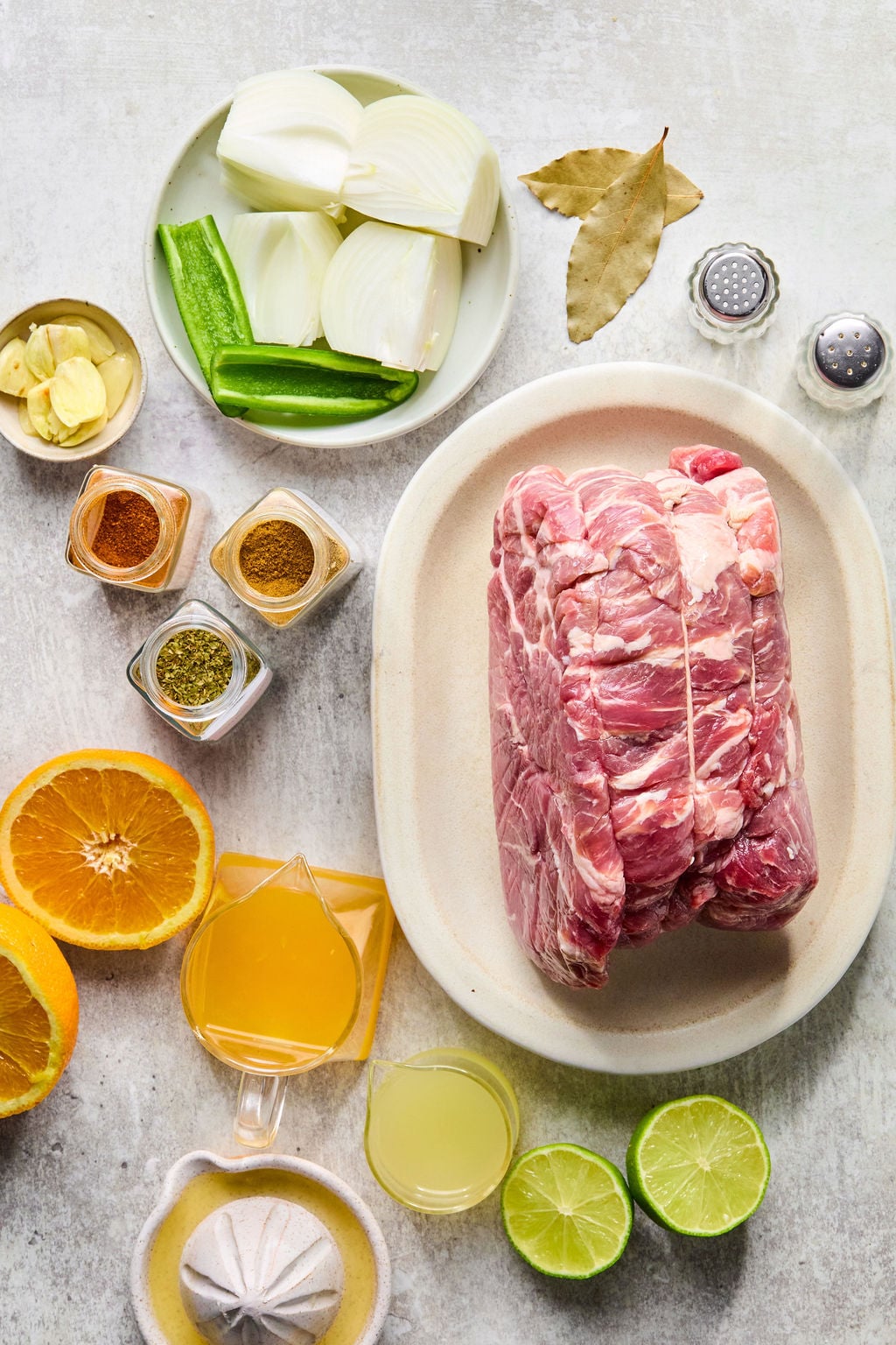 Raw pork roast on a plate surrounded by cut onions, garlic, bay leaves, spices, green chili, halved orange and lime, orange juice, and lime juice on a light surface.