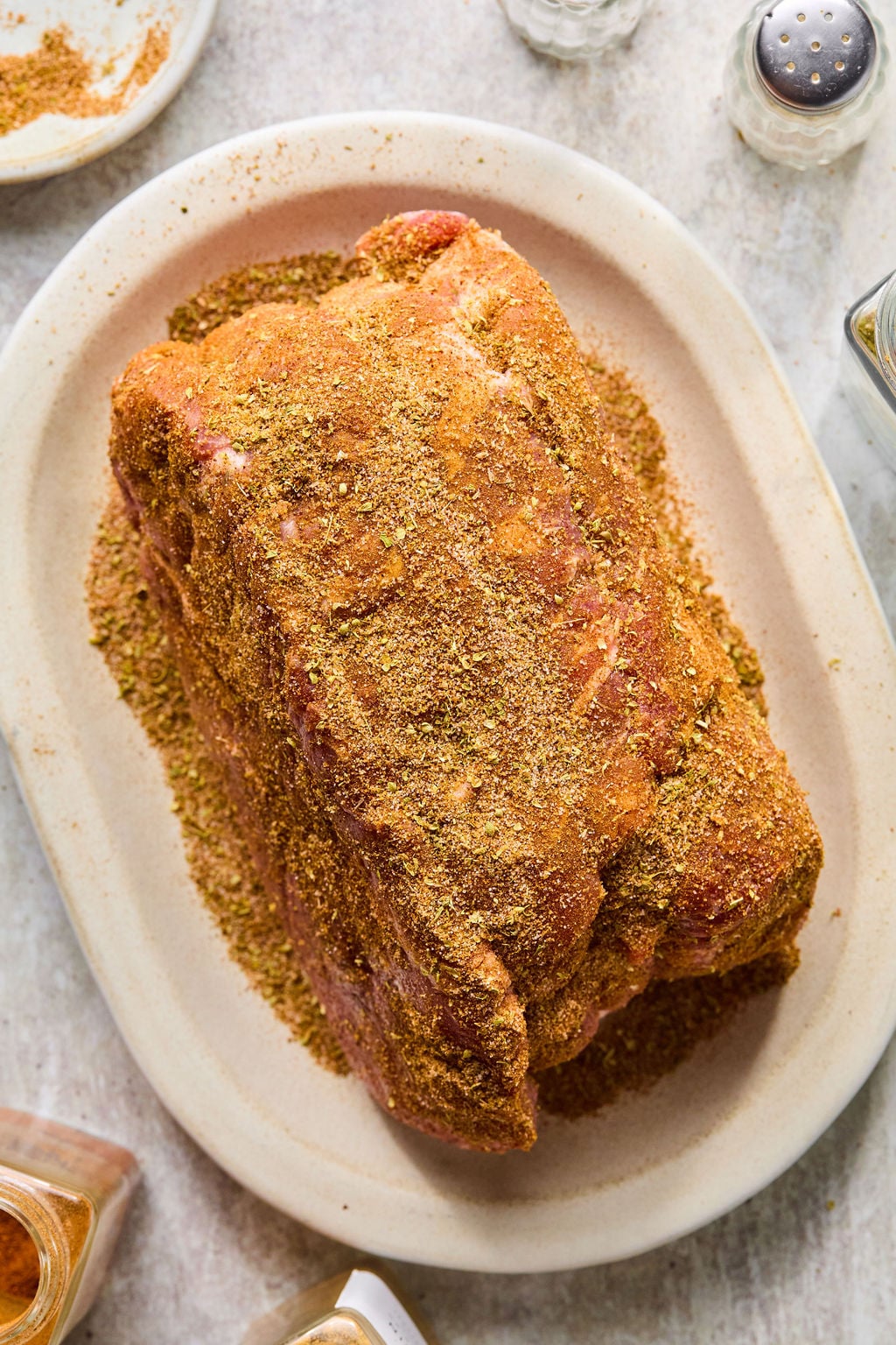 A large piece of raw meat coated with dry seasoning sits on an oval plate, surrounded by spice jars and salt and pepper shakers.