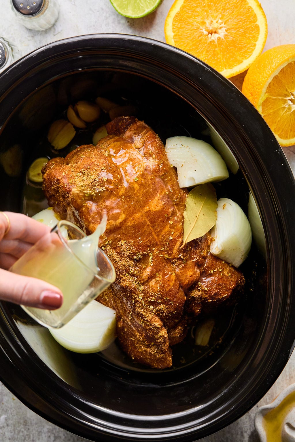 A hand pours liquid over a seasoned pork roast in a slow cooker with onions, garlic, and bay leaves; orange and lime halves are nearby.