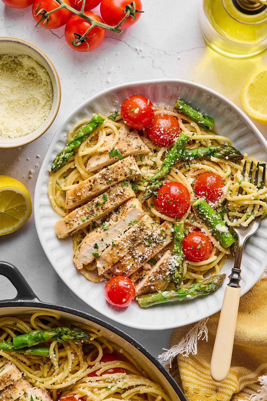 A plate of pasta with grilled chicken slices, asparagus, cherry tomatoes, grated cheese, and herbs, next to a fork, lemon halves, a bowl of cheese, and a bunch of tomatoes.