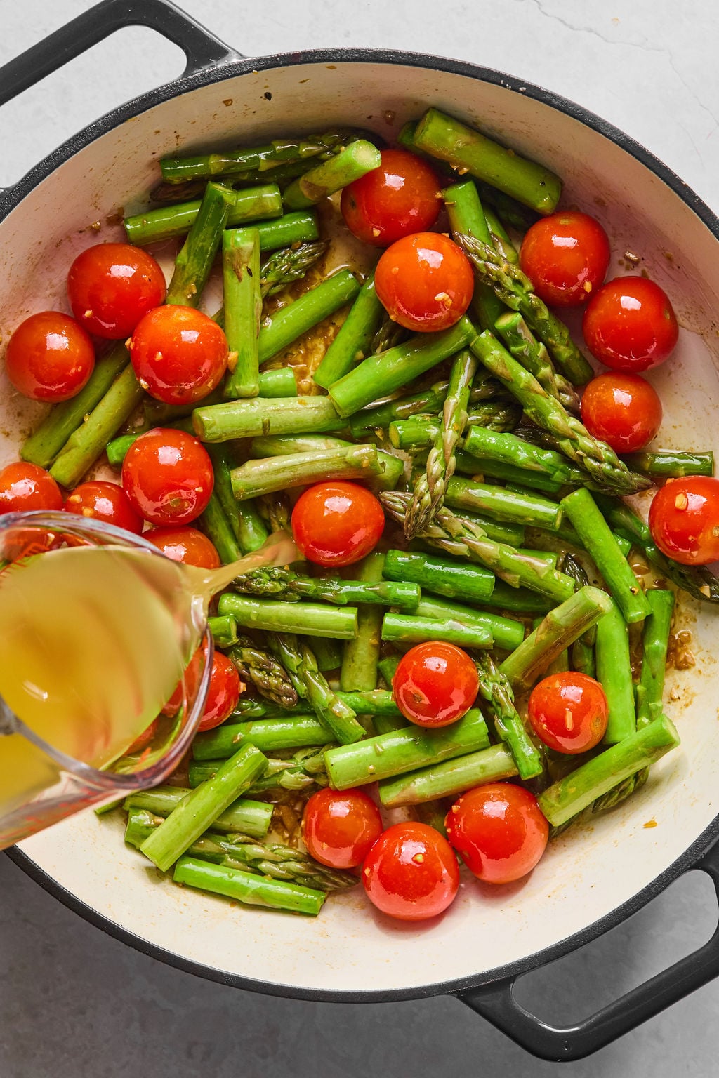 A pot containing asparagus and cherry tomatoes is being cooked, while a clear liquid is being poured into the mixture.