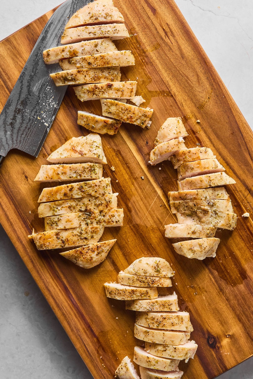 Sliced cooked chicken breast seasoned with herbs on a wooden cutting board, with a knife placed next to the meat.