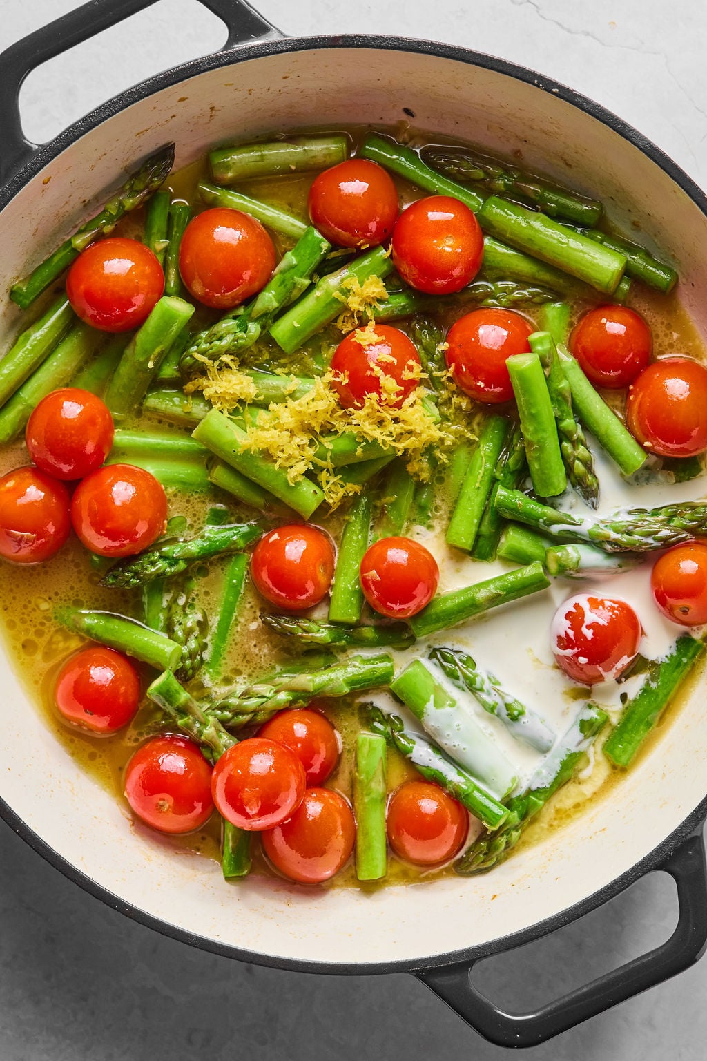 A pan filled with cherry tomatoes, asparagus, lemon zest, and a creamy sauce, all being cooked together.