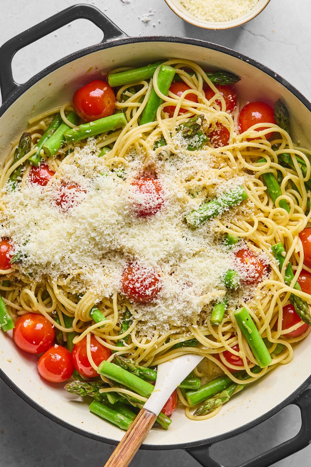 Spaghetti with cherry tomatoes, asparagus, and grated parmesan cheese in a large pan, with a wooden spoon resting inside.