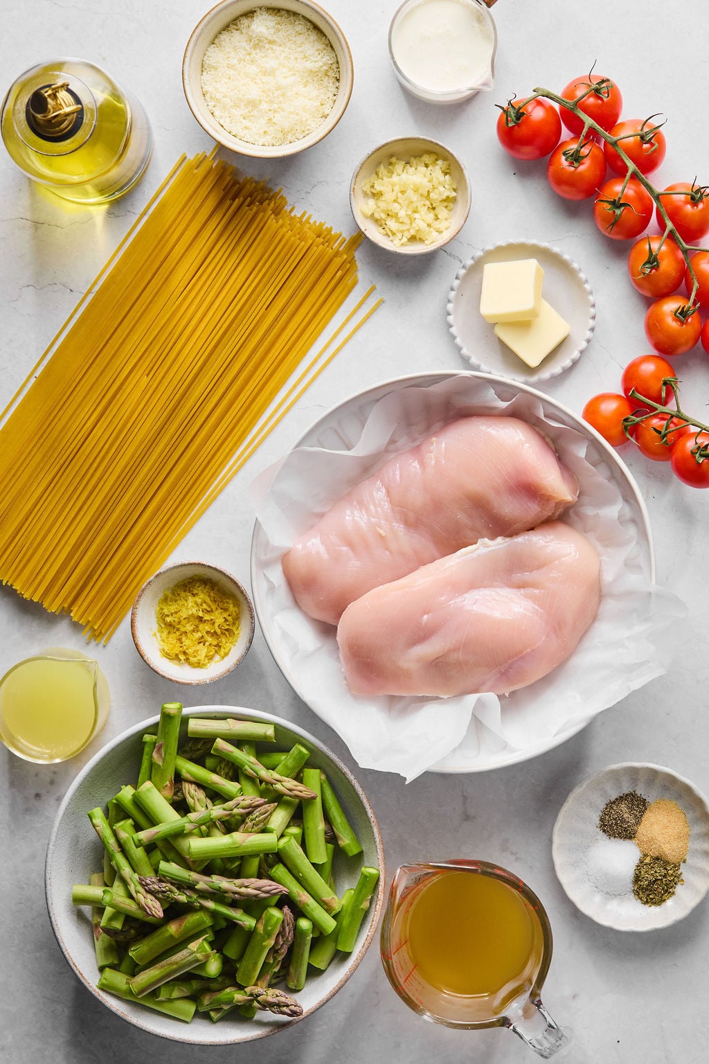 Raw chicken breasts, uncooked pasta, asparagus, cherry tomatoes, butter, grated cheese, cream, oil, minced garlic, lemon zest, chicken broth, and seasonings arranged on a countertop.