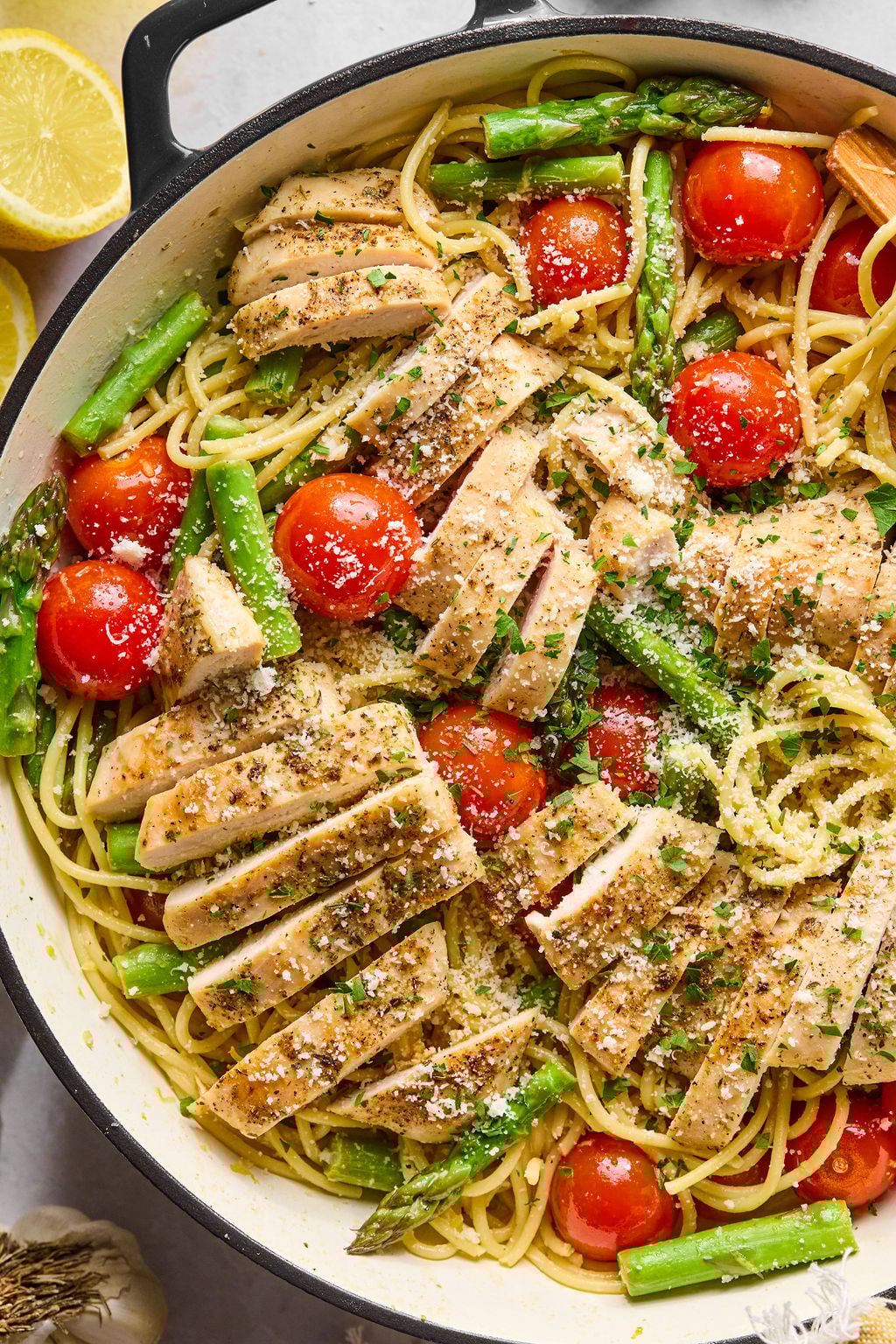 A pan filled with spaghetti topped with sliced grilled chicken, cherry tomatoes, asparagus, grated cheese, and herbs.