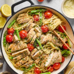 A skillet filled with pasta, sliced grilled chicken, asparagus, cherry tomatoes, and grated cheese, surrounded by fresh tomatoes, lemon, garlic, olive oil, and a bowl of grated cheese.