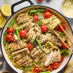 A skillet filled with pasta, sliced grilled chicken, asparagus, cherry tomatoes, and grated cheese, surrounded by fresh tomatoes, lemon, garlic, olive oil, and a bowl of grated cheese.