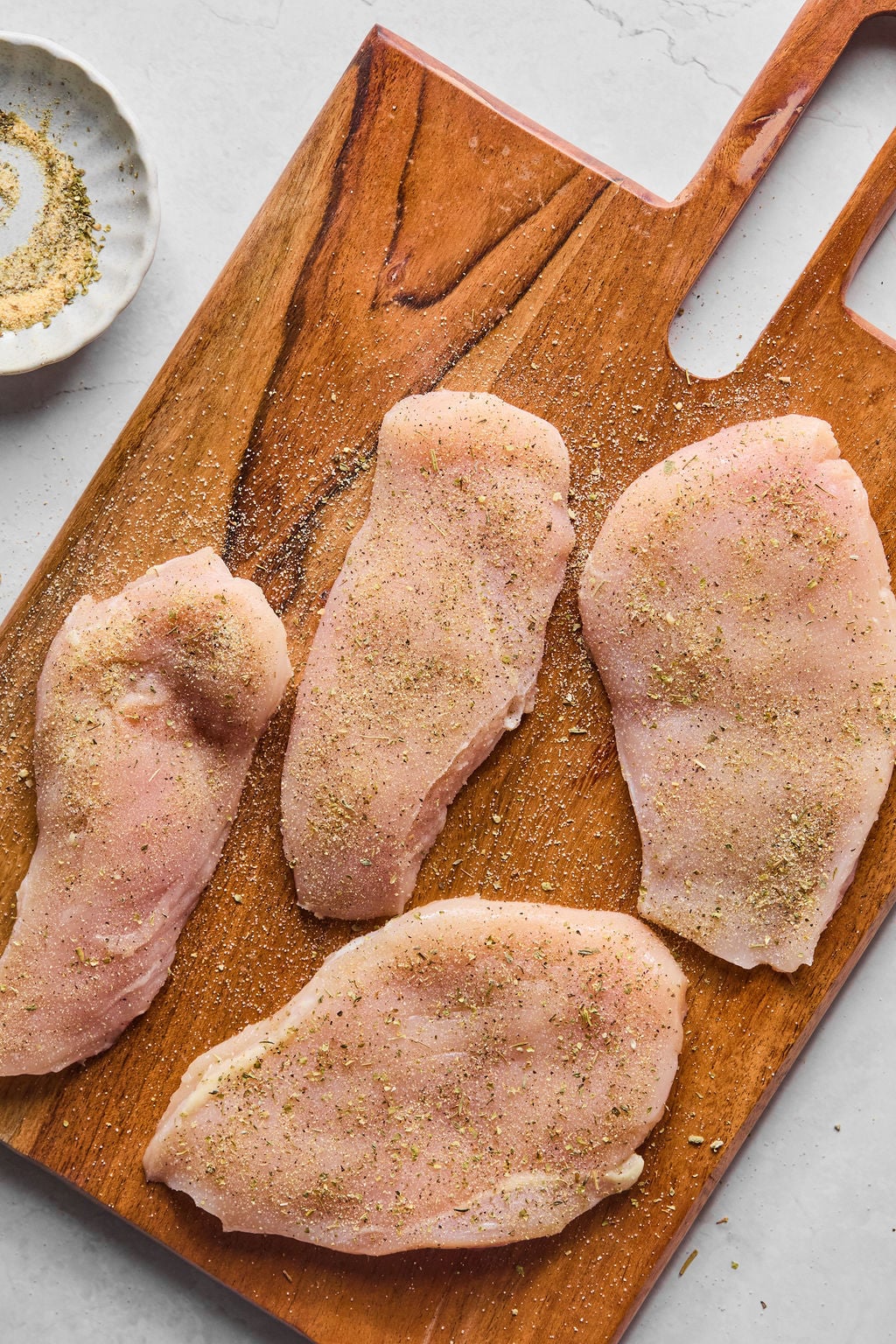 Four raw chicken breasts seasoned with spices are arranged on a wooden cutting board, with a small dish of seasoning nearby.