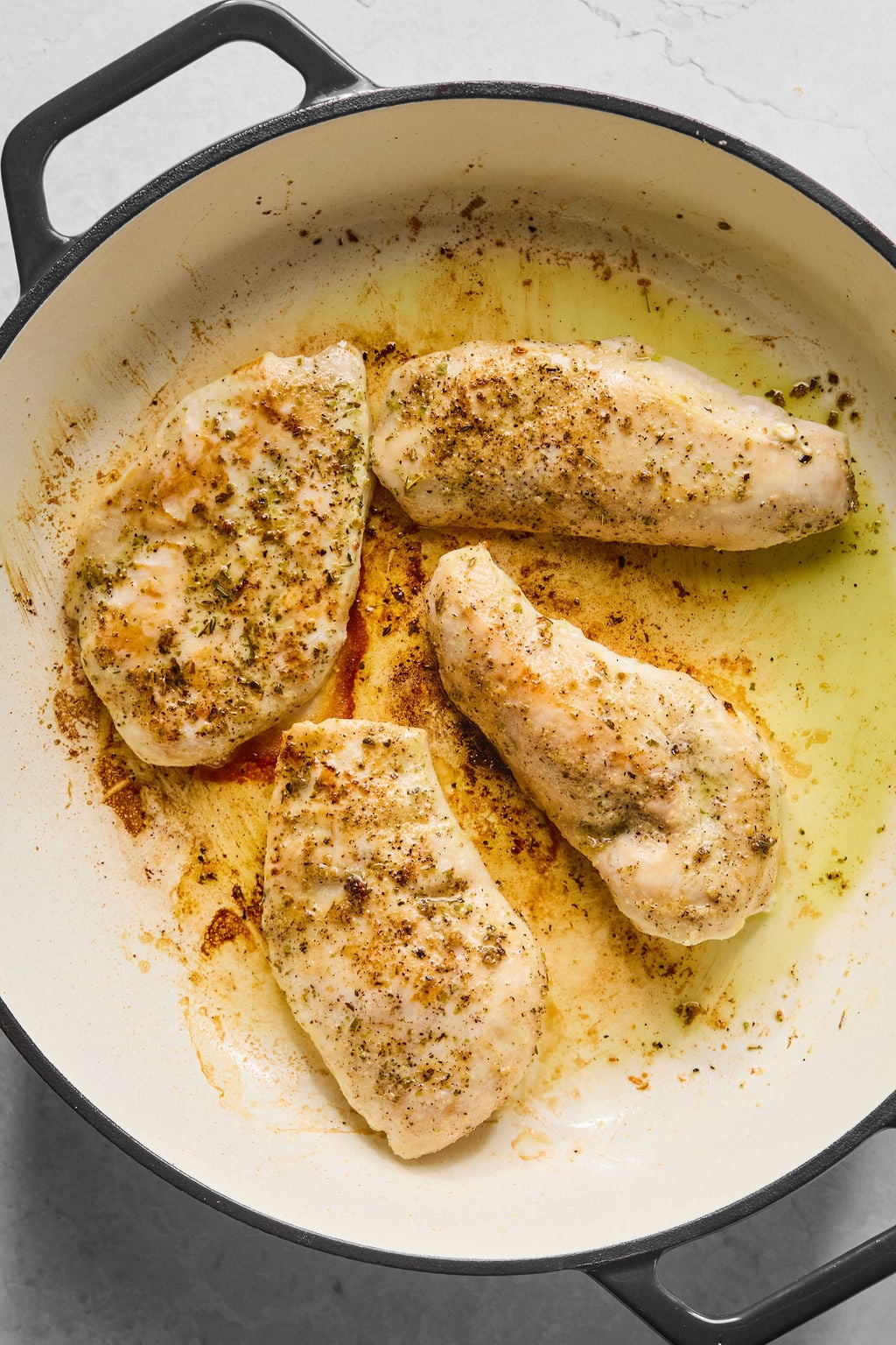 Four seasoned chicken breasts cooking in a large white skillet with some browned residue and oil visible on the pan.