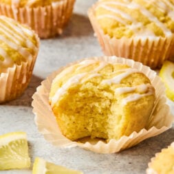 Lemon muffins with icing drizzled on top, one muffin partially eaten, surrounded by lemon slices on a light surface.