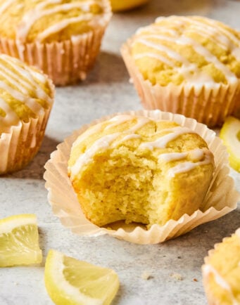 Lemon muffins with icing drizzled on top, one muffin partially eaten, surrounded by lemon slices on a light surface.