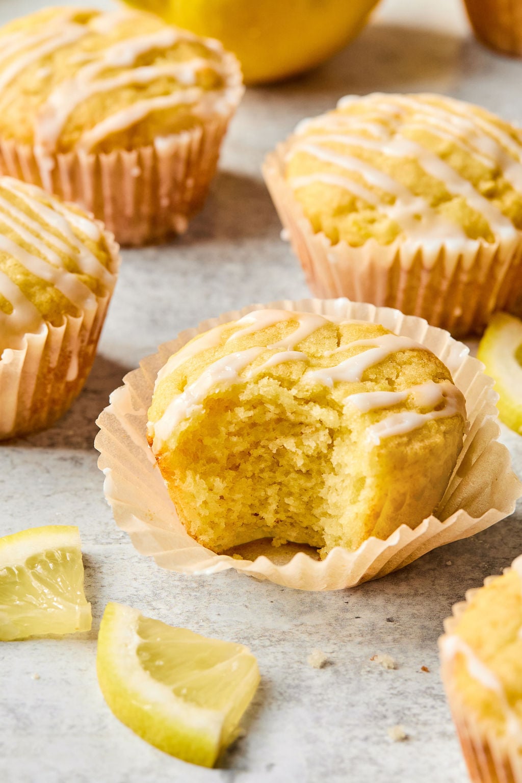 Lemon muffins with icing drizzled on top, one muffin partially eaten, surrounded by lemon slices on a light surface.