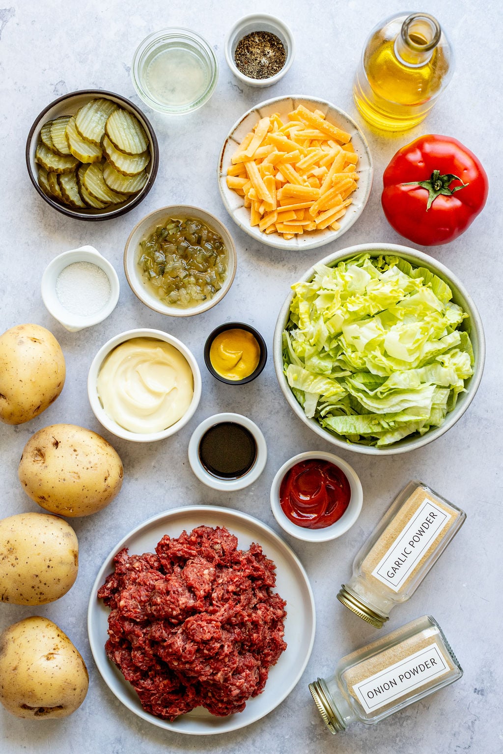 Overhead view of ingredients including ground beef, potatoes, lettuce, tomato, cheddar cheese, pickles, condiments, spices, and oil arranged on a light surface.