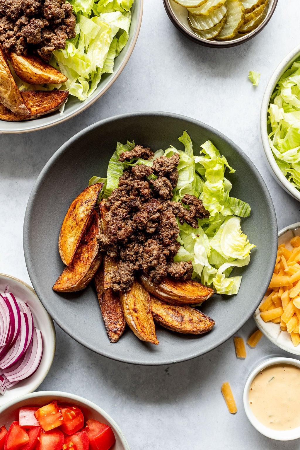 A bowl with seasoned ground beef, lettuce, and potato wedges, surrounded by bowls of sliced pickles, shredded cheese, chopped lettuce, sliced onions, diced tomatoes, and dressing.