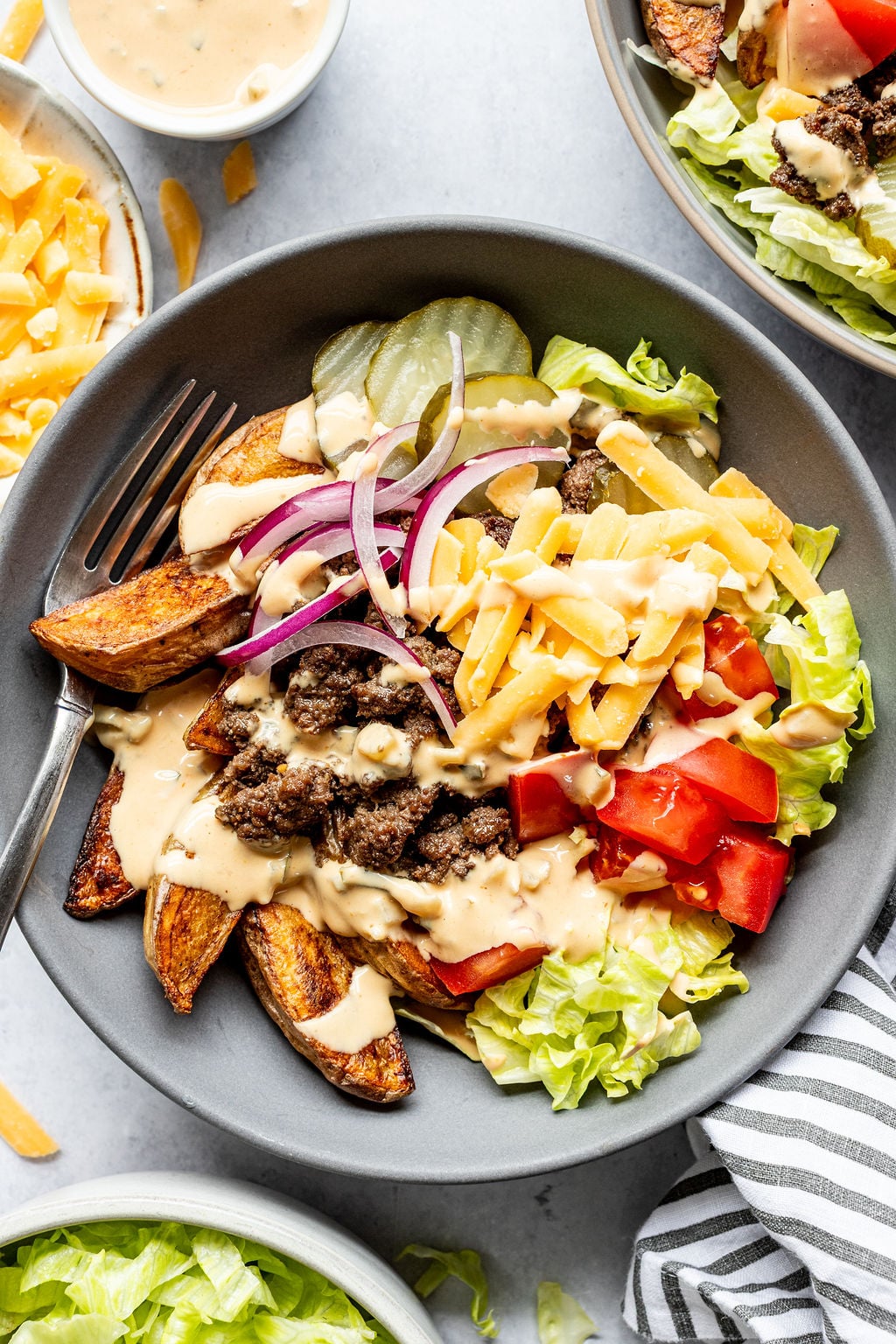 A bowl of lettuce, tomato, shredded cheddar cheese, ground beef, pickles, potato wedges, and sliced red onion, topped with a creamy sauce.
