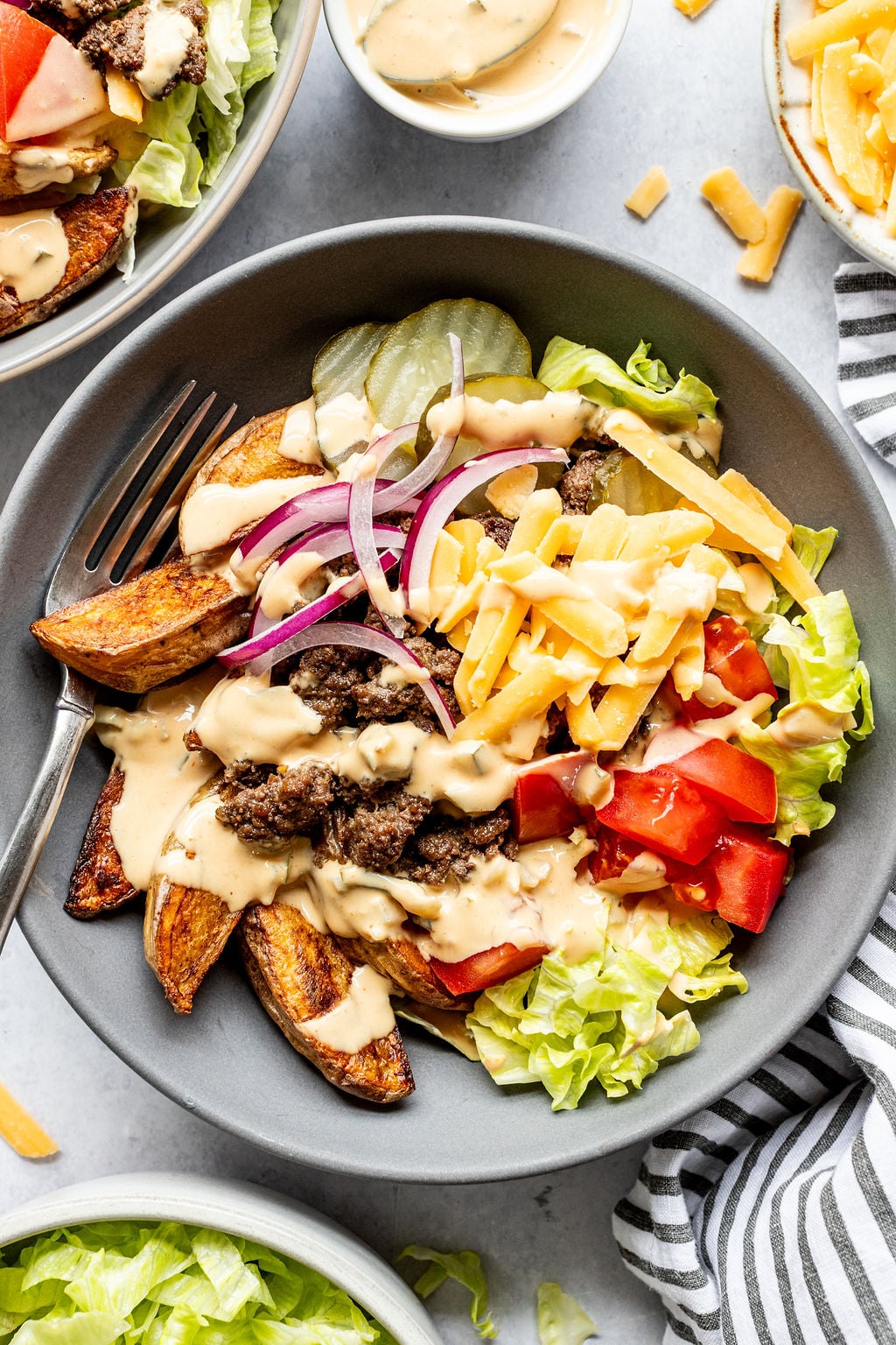 A bowl filled with lettuce, tomato, cheddar cheese, red onion, pickles, beef, potato wedges, and creamy dressing, with a fork on the side.
